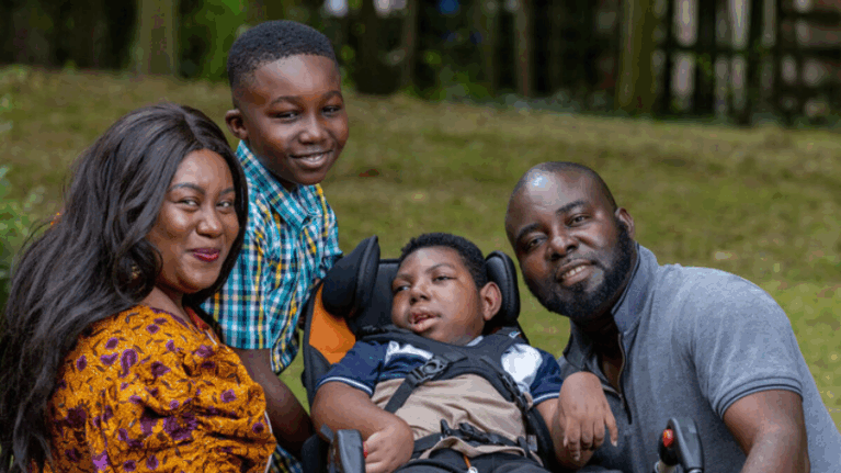 A family of four smile at the camera in a park. Left to right are mum in a yellow dress, a young son in a blue shirt, his brother reclining in a wheelchair and their dad.