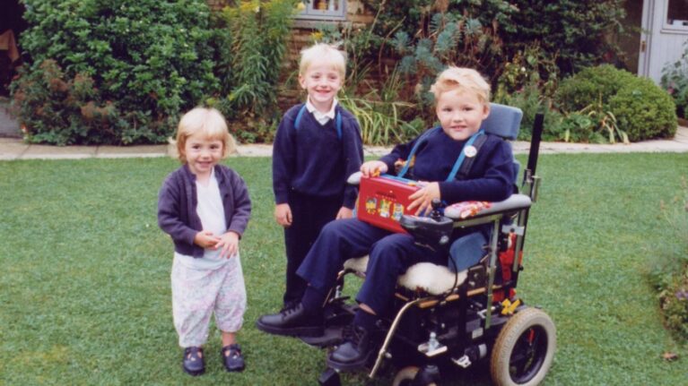 Three children smile at the camera. (L-R) a young girl stands next to an older boy. Next to him is a young boy in a wheelchair.
