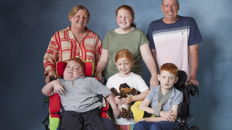 A family of five pose for a photo. In the back row are mum, daughter and dad. In between two sons in wheelchairs in the front row is another daughter holding a teddy.