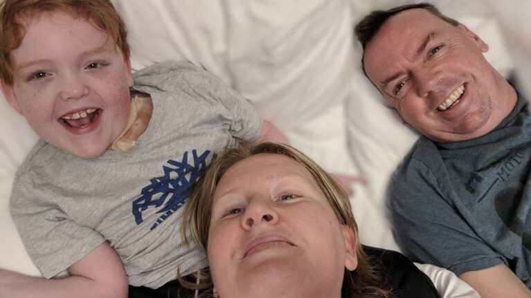 A family of three pose for a selfie in bed, all three are smiling at the camera.