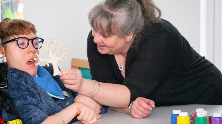 A mum in a black top is holding a paper cut-out of a tree to her son who is in a wheelchair. In front of them is an arts and crafts table.