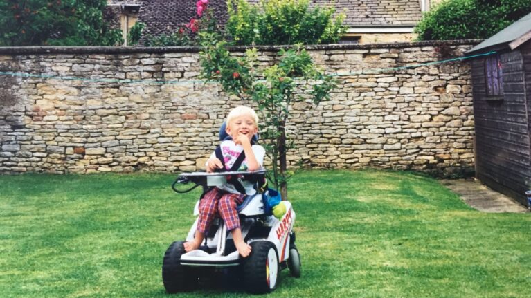 A boy sits in his wheelchair in the garden. Behind him is a high stone wall.