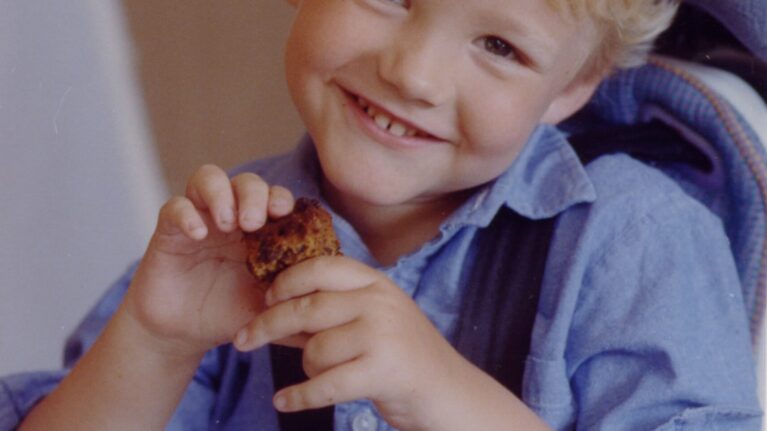 A blonde-haired boy smiles at the camera. Wearing a blue shirt, he is holding his hands together in front of himself.