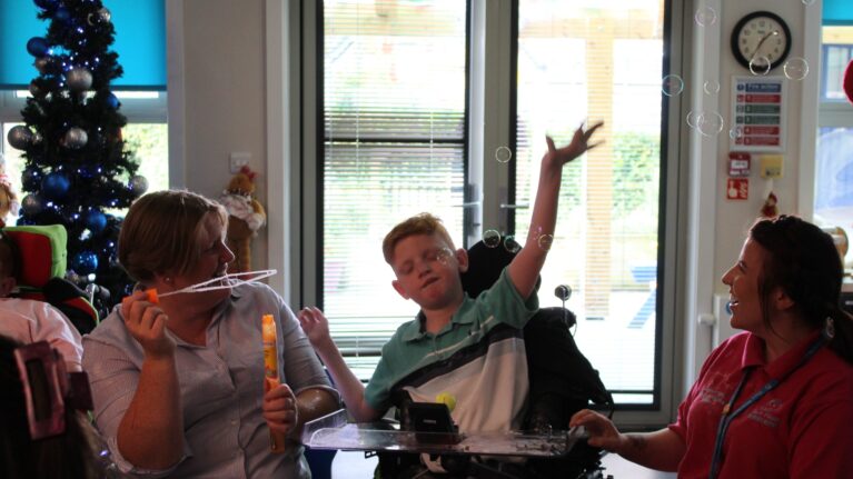 A young boy in a wheelchair is waving his hands as his two smiling carers blow bubbles around him