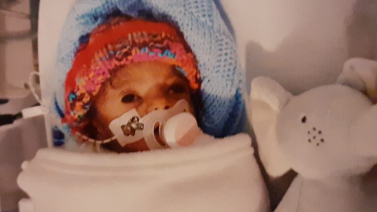 A premature baby is wrapped up in blankets in an incubator. He is wearing an orange woolly hat and has a dummy in his mouth. A Hickman line with a teddy bear plaster is visible and next to him is a cuddly elephant toy.