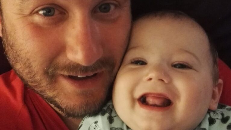 A dad in a red top is smiling at the camera. In his arms and giggling is a baby boy wearing a mickey mouse onesie.