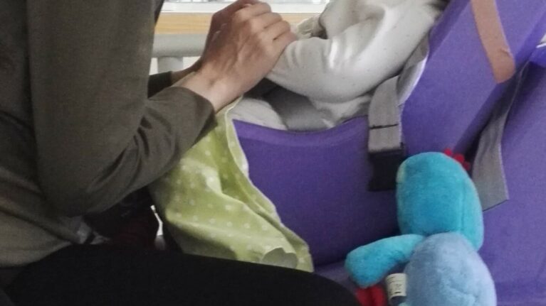 A baby boy in a purple baby carrier is holding hands with his mum; they are smiling at each other. A Hickman Line can be seen on the little boys face.