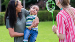 A young boy is being held in his mothers arms in a garden. A lady stands in front of them blowing bubbles