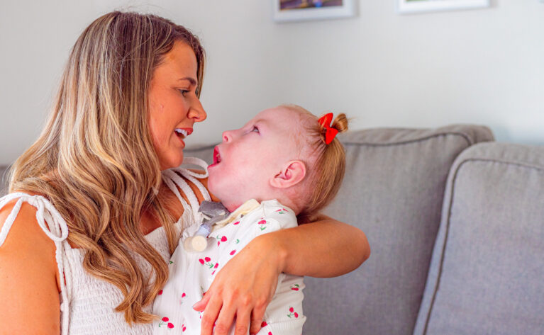 A mum is sat on the sofa cradling her daughter. They are both gazing into each others eyes
