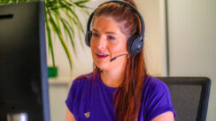 a white woman with long brown hair tied back and wearing a purple t-shirt is talking through a headset. she is sat at a desk