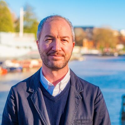 A man smiles at the camera. He is wearing a blue suit. Behind him is Bristol harbourside