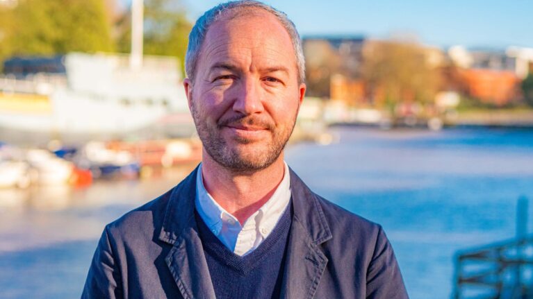 A man smiles at the camera. He is wearing a blue suit. Behind him is Bristol harbourside