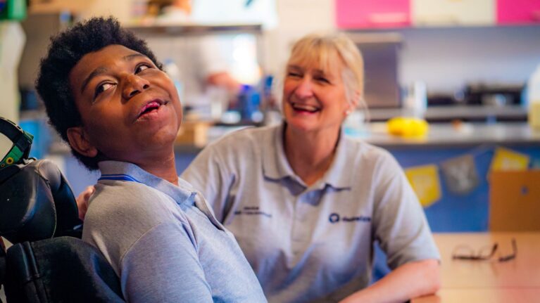 A young person sat in a wheelchair is smiling and looking at something behind the camera. A care professional sitting with him is also smiling.