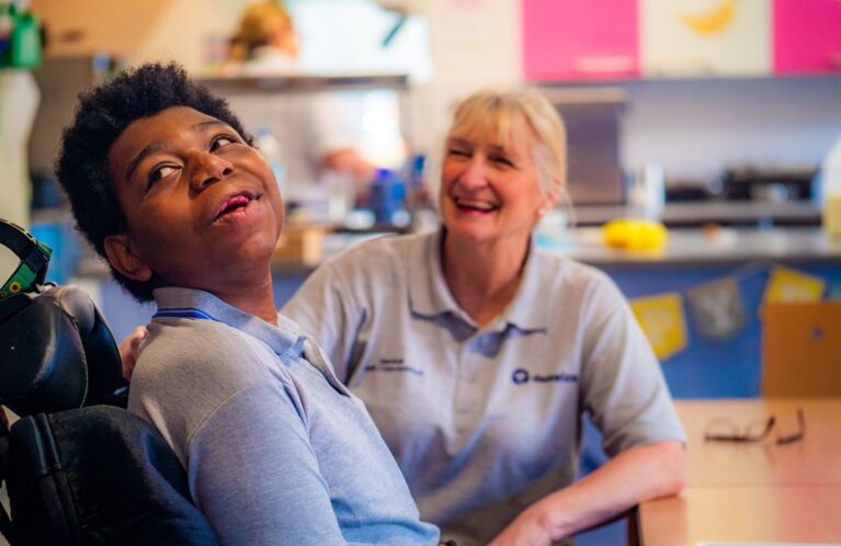 A young person sat in a wheelchair is smiling and looking at something behind the camera. A care professional sitting with him is also smiling.