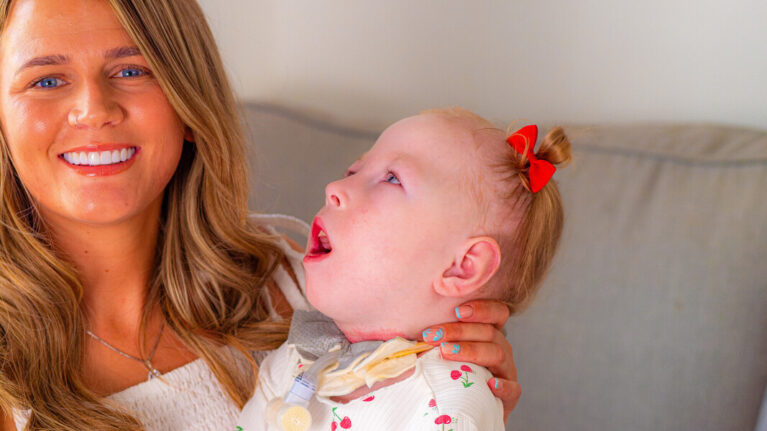 A white lady with blonde hair smiles at the camera. She is hugging her blonde baby who has red ribbons in her hair