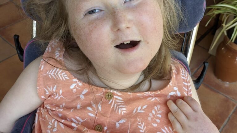 A young girl in a wheelchair is smiling up at the camera